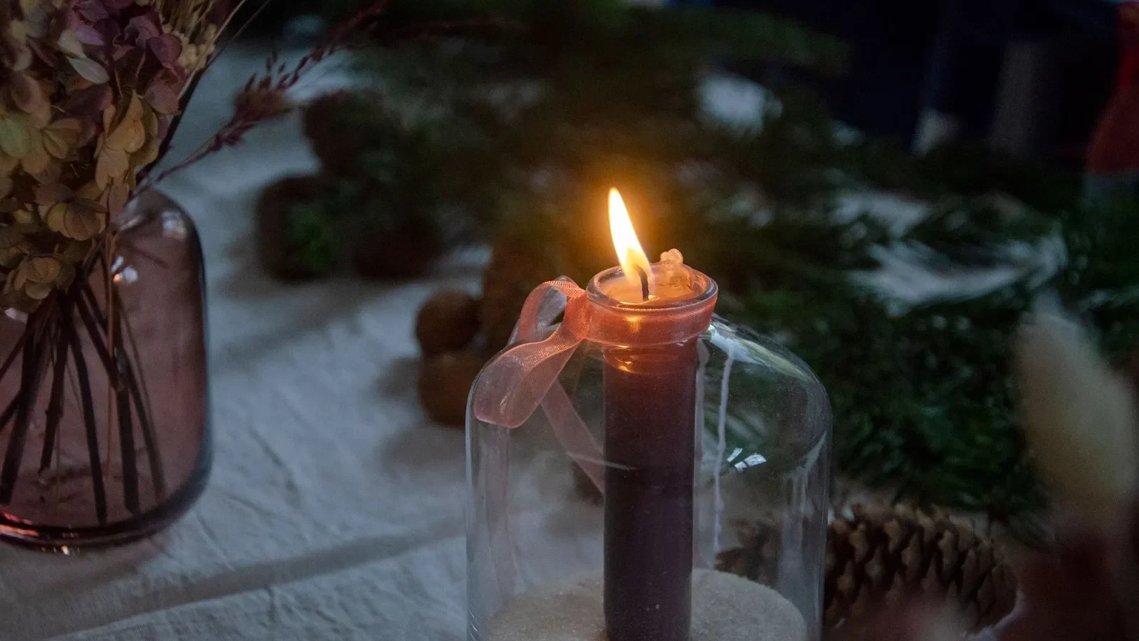 Bougie allumée dans un contenant en verre entourée de fleurs et de pommes de pin