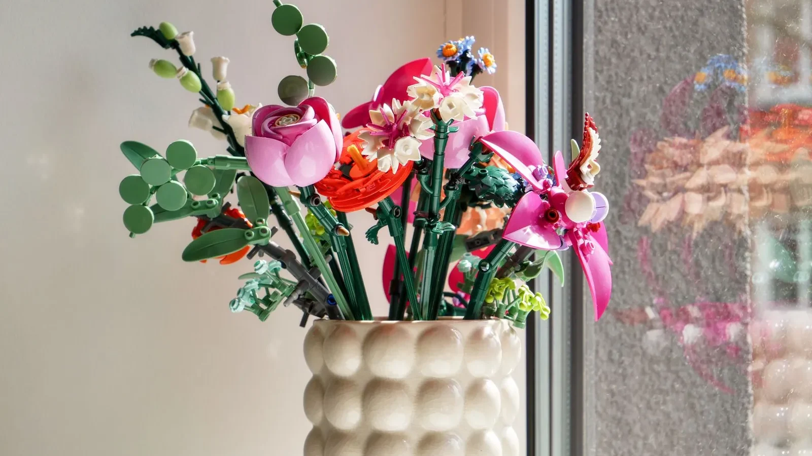 Fleurs artificielles colorées dans un vase texturé près d’une fenêtre